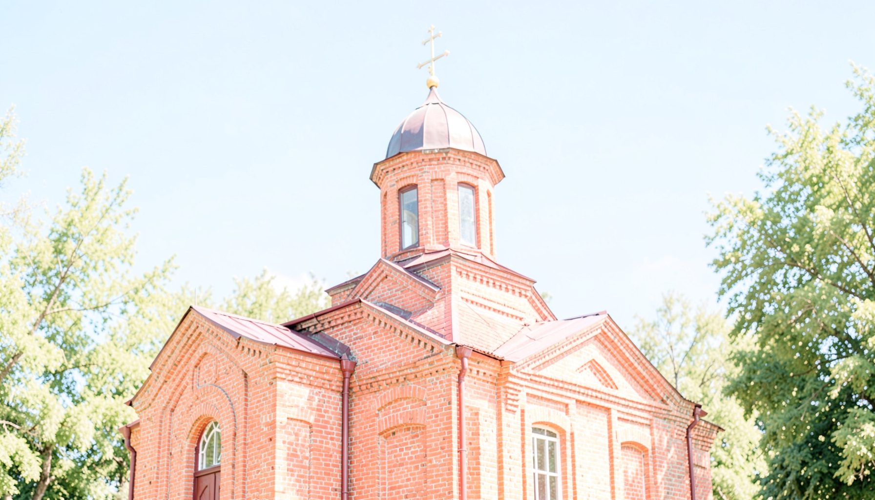 A bright, airy photograph of a modest Orthodox church building with a red brick facade and a cross-topped dome, set against a clear blue sky with soft green trees in the background, conveying a peaceful and welcoming atmosphere.