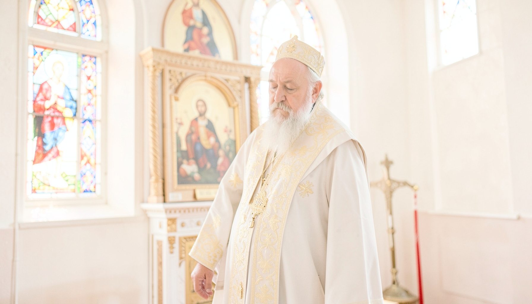 A bright, airy photograph of an Orthodox priest in vestments standing before an iconostasis, with soft daylight filtering through stained glass.