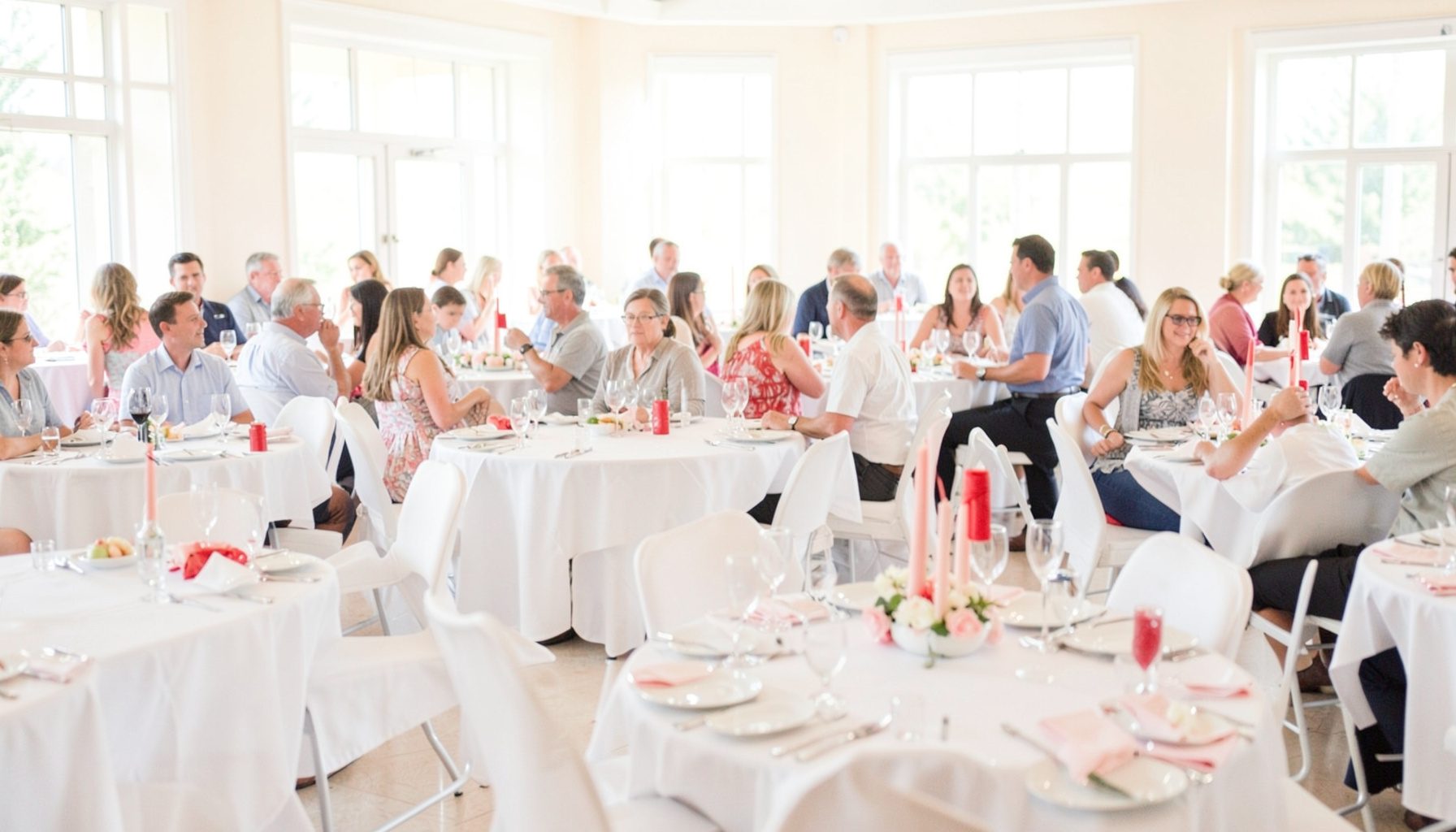 A bright, airy photograph of a community gathering in a banquet hall with round tables, white tablecloths, and soft natural light from large windows.
