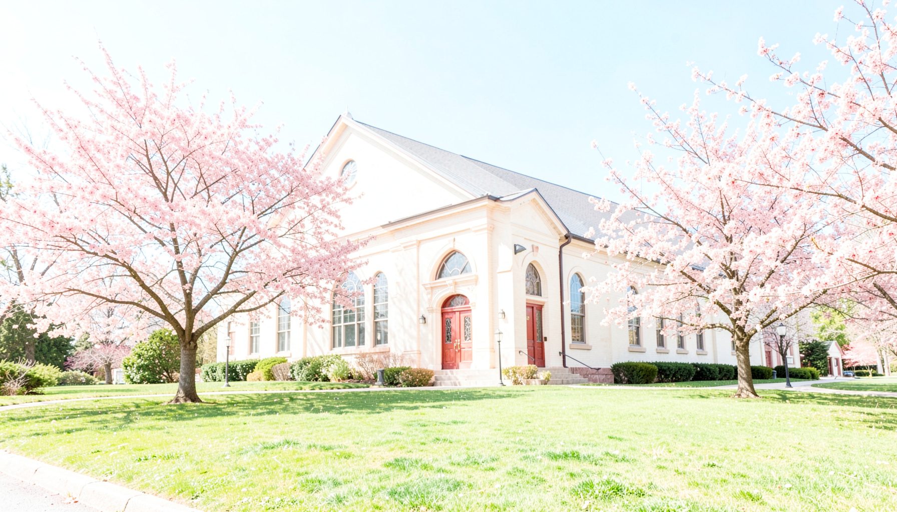A bright, airy photograph of the church exterior in springtime, with flowering trees and a well-kept lawn under a gentle blue sky.