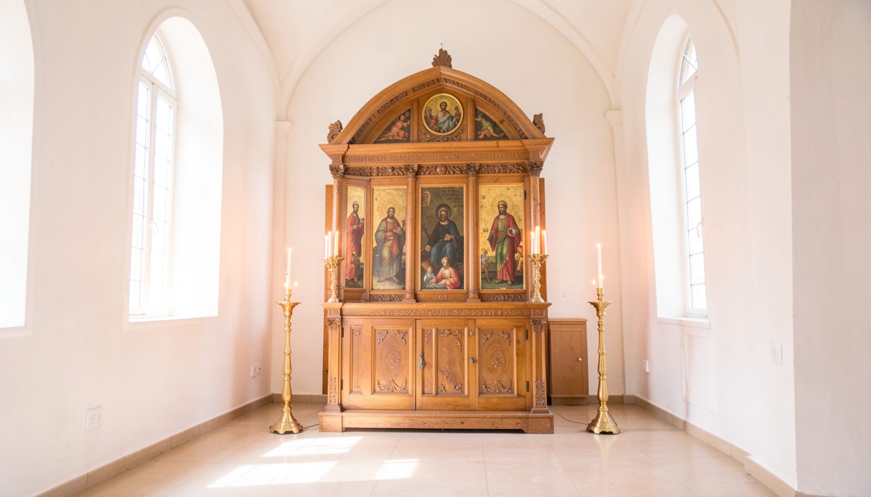 Sunlit interior of a serene Orthodox church with warm wooden iconostasis, glowing candle stands, and soft light filtering through arched windows