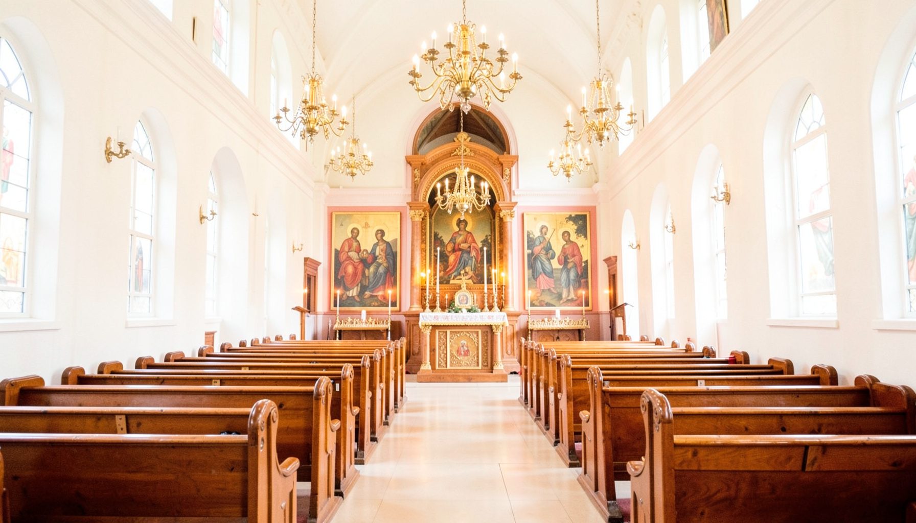 Warm interior view of an Orthodox church nave with polished wooden pews, hanging brass chandeliers, and an iconostasis glowing in candlelight