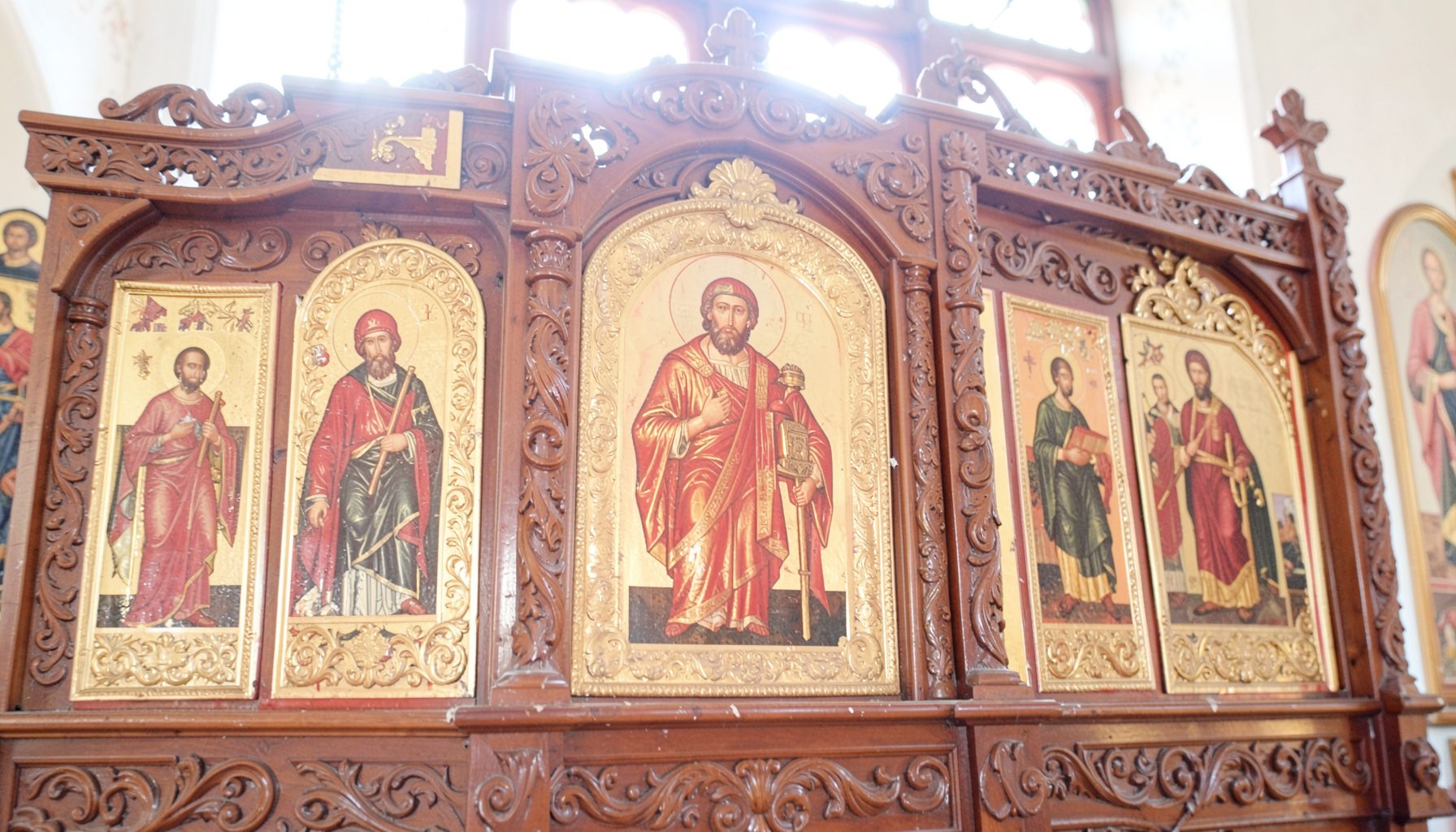 Close-up of ornate Orthodox icons in gold and deep red tones, mounted on a carved wooden iconostasis with soft natural light from above