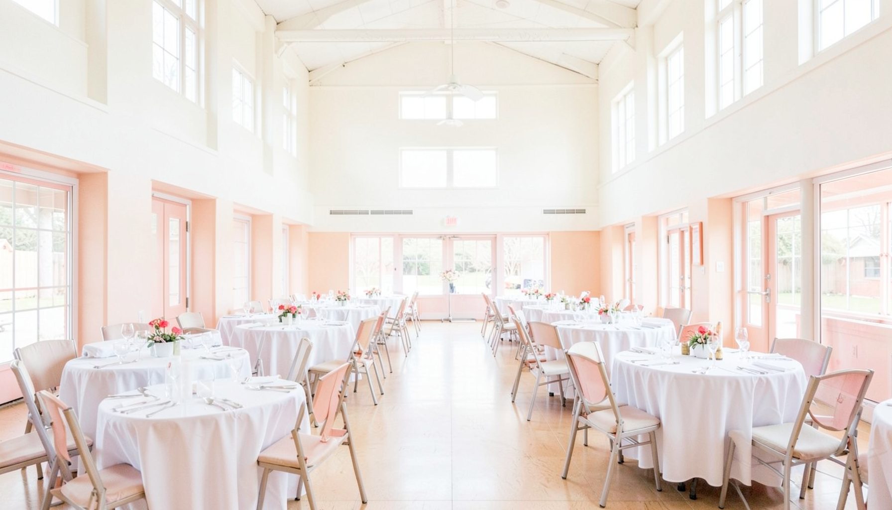 Bright community hall with rows of round tables set with white linens, high ceiling, and large windows letting in daylight