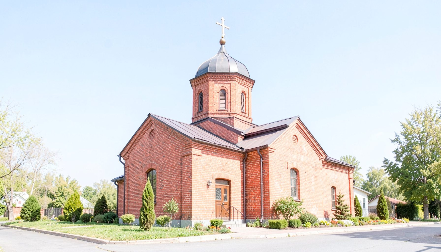 Exterior of a modest brick Orthodox church under a clear blue sky, with a cross-topped dome and tidy landscaping