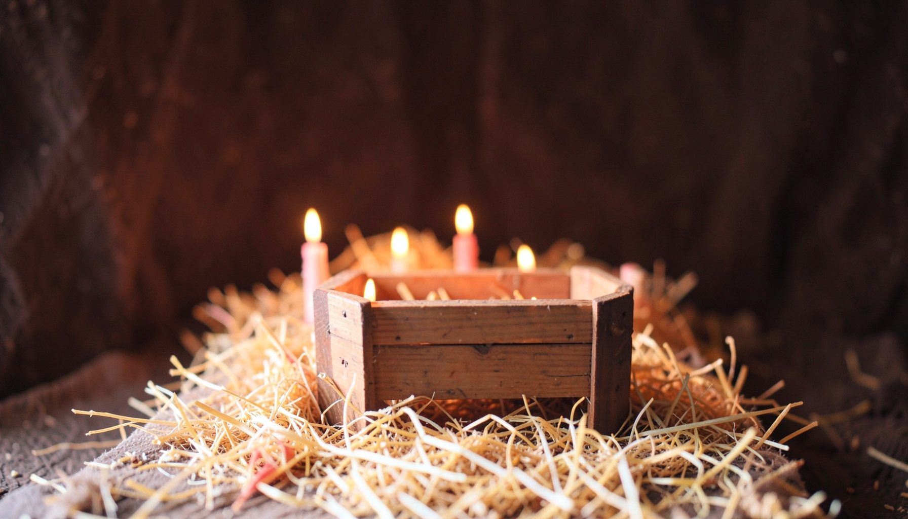 A softly lit nativity scene with a wooden manger on straw, warm candlelight glowing against a dark background, deep browns and golds
