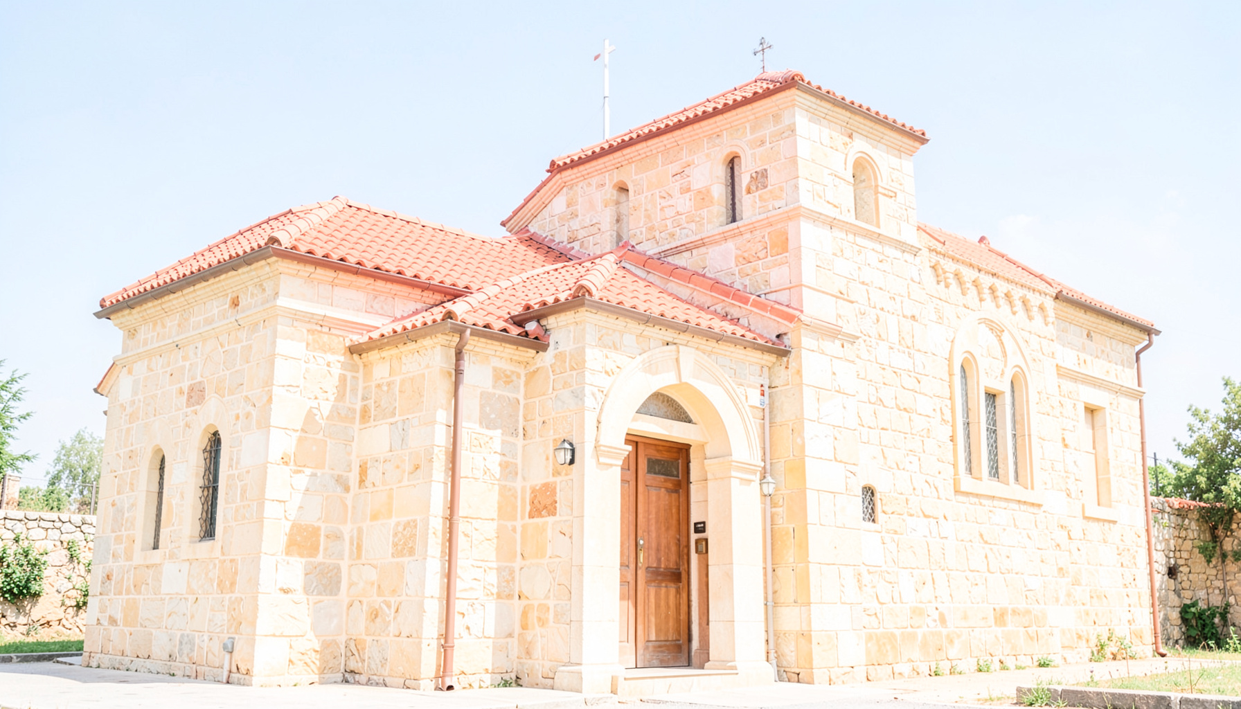 Bright, airy photograph of a modest Orthodox church exterior with warm beige stone walls and a red-tiled roof, set against a clear blue sky with soft sunlight casting gentle shadows across the front entrance.